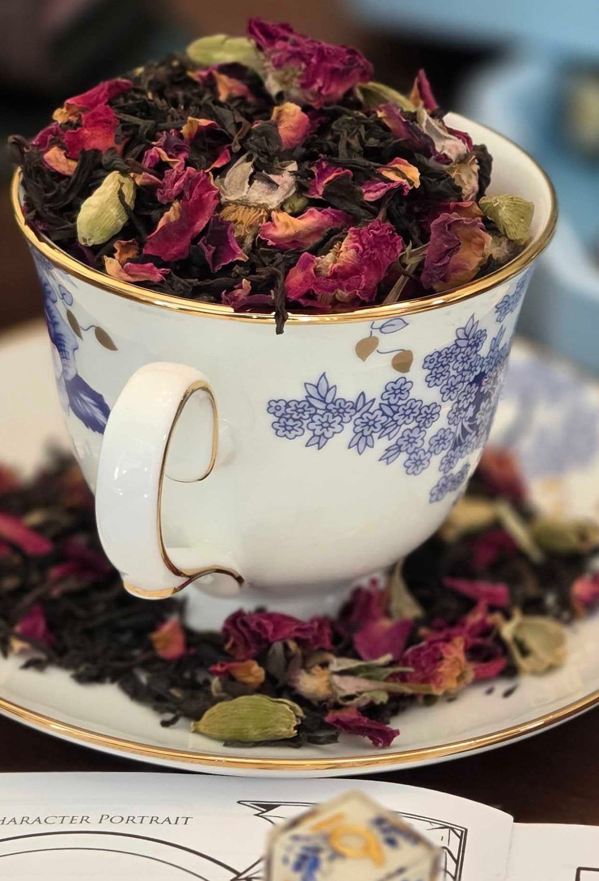 Teacup filled with dried flowers on a saucer, surrounded by books and dice on a table.