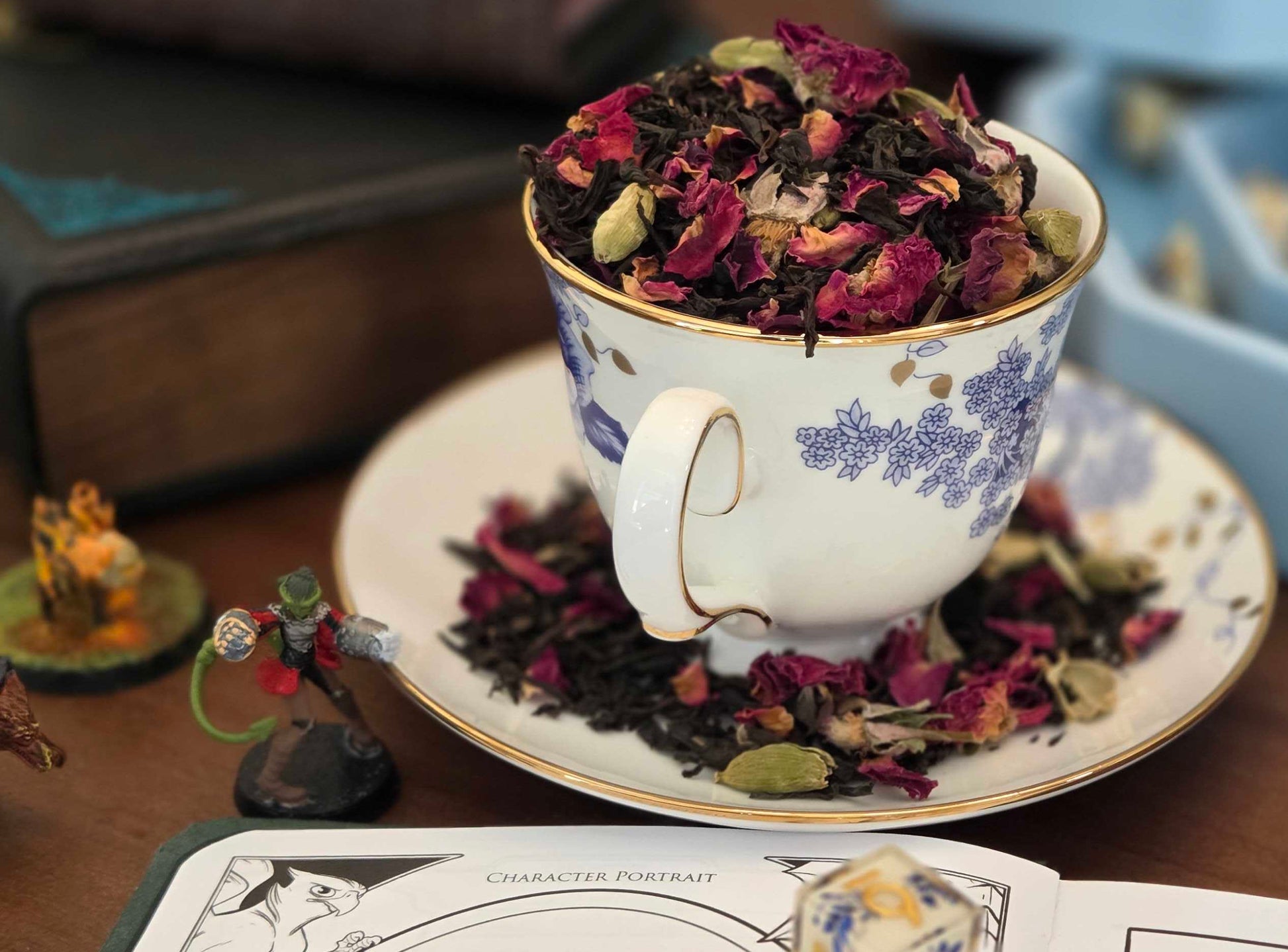 Teacup filled with dried flowers on a saucer, surrounded by books and dice on a table.